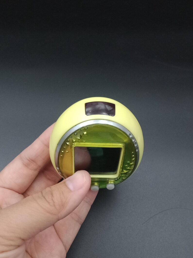 May include: A handheld, yellow electronic device with a rectangular screen and two gray buttons. The device has a silver trim and a small, dark screen at the top. The device is held in a hand against a dark background.