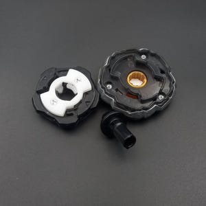 Pu&ograve; includere: Un trottola giocattolo nera e argento smontata con un componente bianco e nero. La trottola ha un'impugnatura nera e un anello esterno argentato. I componenti sono disposti su una superficie grigio scuro.