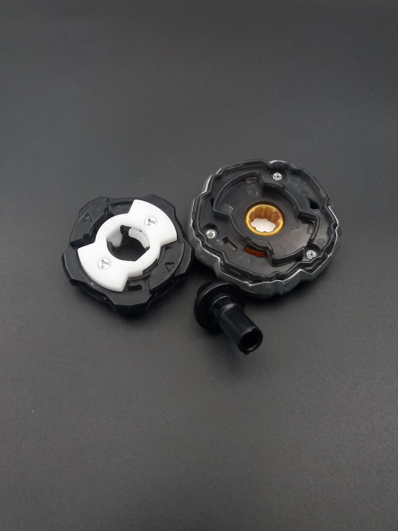 Pu&ograve; includere: Un trottola giocattolo nera e argento smontata con un componente bianco e nero. La trottola ha un'impugnatura nera e un anello esterno argentato. I componenti sono disposti su una superficie grigio scuro.