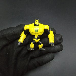 May include: A yellow action figure with black detailing and a green emblem on its chest. The figure has a muscular build and is posed with arms at its sides. The toy is held in a black gloved hand against a dark background.