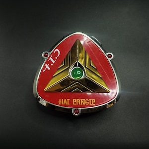 May include: A triangular, metallic badge with a red border and gold center. The center features a green gem and the text "+I.T." and "HAI PANGIE" in gold lettering. The badge has a silver trim and is set against a dark background.