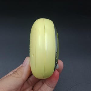 May include: A pale yellow, oval-shaped electronic device with a silver trim. The device is held in a hand, and the side profile shows a small button and a seam. The background is a dark gray.