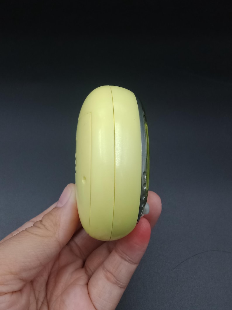 May include: A pale yellow, oval-shaped electronic device with a silver trim. The device is held in a hand, and the side profile shows a small button and a seam. The background is a dark gray.