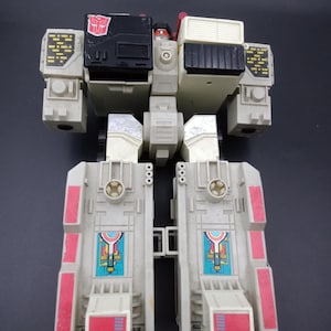 May include: A vintage toy robot in shades of gray, black, and red. The toy has a blocky design with detailed panels, including a red and blue emblem. The robot's legs are split, and it has a rectangular torso.