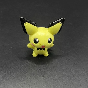 Puede incluir: Una figura amarilla de un personaje de dibujos animados con grandes orejas con puntas negras, ojos negros y una boca pequeña. El personaje tiene brazos pequeños y una pajarita negra. La figura está sobre una superficie oscura.