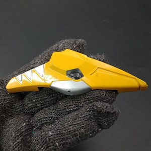 Pode incluir: Um componente de plástico amarelo e prateado com um design geométrico. A peça tem uma forma triangular com uma secção prateada e um detalhe preto. O item é segurado por uma luva cinza escura.