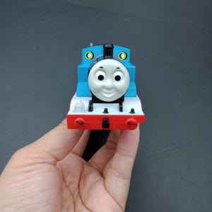 May include: A blue, red, and white toy train with a smiling face. The toy train has a black smokestack and yellow circular details. The train is held in a person's hand against a dark background.
