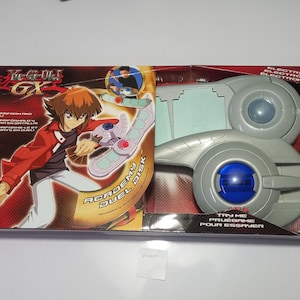 Peut inclure: Un jouet électronique Yu-Gi-Oh! GX Academy Duel Disk dans son emballage rectangulaire d'origine. La boîte présente une illustration d'un personnage portant l'appareil en plastique gris, doté d'un centre bleu circulaire et d'un emplacement pour cartes.