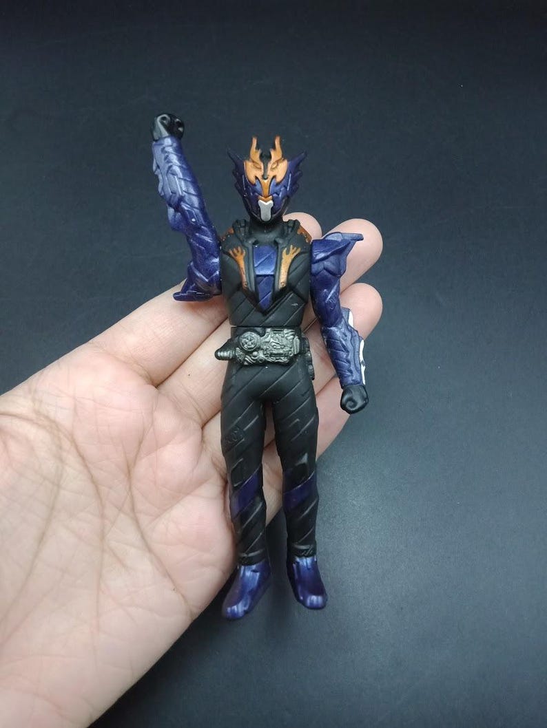 May include: A detailed action figure of a character in a black and blue suit with a silver belt. The figure has a helmet with orange accents and a raised arm. The figure is approximately 6 inches tall.