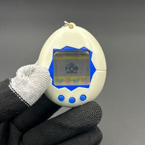 May include: A vintage egg-shaped digital pet toy with a small screen displaying pixelated graphics. The toy is white with blue accents and three blue buttons. A gold-colored chain is attached to the top.