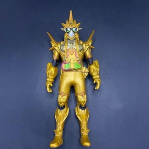 Puede incluir: Figura de acción dorada con diseño detallado. La figura tiene un casco con una espiga central y múltiples protuberancias angulares. Presenta detalles verdes y azules en la cara y un cinturón verde y rosa. La figura está sobre una superficie azul oscuro.