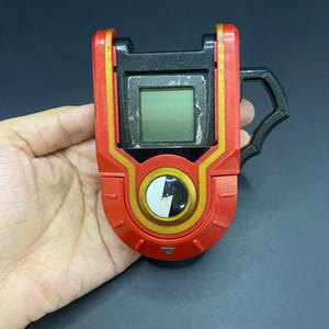Puede incluir: Un dispositivo electrónico portátil rojo y negro con una pequeña pantalla y un botón circular. El dispositivo tiene detalles dorados y un asa en el lateral. La pantalla es un pequeño rectángulo. El dispositivo se sostiene en una mano.