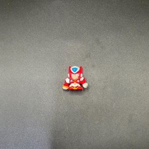 May include: A small, red and white figurine of a character with a blue helmet and accents. The figure is in a seated position, with details including a chest plate and boots. The background is a solid, dark gray.
