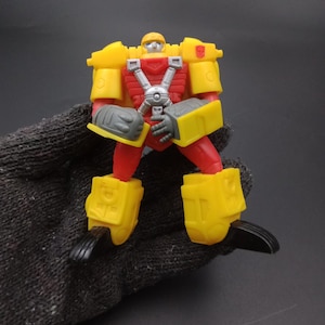 May include: A yellow and red toy robot with gray and black accents. The robot has a helmet, chest plate, and large shoulder pads. The robot's arms and legs are articulated. The toy is a collectible item.