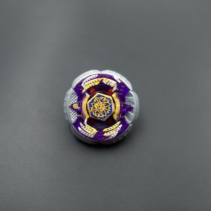 Könnte beinhalten: Ein kreisförmiges Kreiselspielzeug mit einem äußeren Ring aus silberfarbenem Metall, einem violetten Kunststoffmittelteil mit gelben Akzenten und einem zentralen sechseckigen Emblem, das einen gelben Löwen auf blauem Grund zeigt.