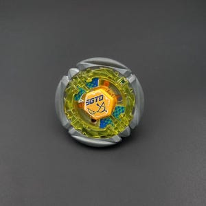 Puede incluir: Un juguete de peonza circular con un anillo exterior metálico plateado, una sección central de plástico amarillo translúcido con patrones de cuadros azules y una tapa hexagonal naranja central con el texto SGTO y un gráfico de ojo estilizado.