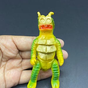May include: A vintage monster toy figurine in yellow and green. The toy has a segmented body, a red mouth, and two small horns. The figure is held in a hand against a dark background.