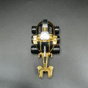 Könnte beinhalten: Ein schwarz-goldenes Spielzeugauto mit sechs schwarzen Rädern. Das Auto hat eine goldene Front und Akzente, mit den Buchstaben "BF" an den Seiten. Das Auto hat ein futuristisches Design.