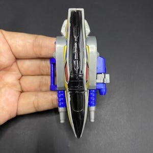 May include: A colourful toy with a black, yellow, and silver body, red accents, and blue and silver details. The toy is held in a hand and appears to be a part of a larger set or figure. The toy has a sleek, aerodynamic design.