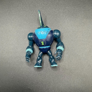 Op de afbeelding: Een blauw en turquoise actiefiguur van een personage uit de animatieserie Ben 10. Het figuur heeft een donkerblauwe body met turquoise accenten op de borst, schouders en voeten. Een groen en zwart embleem staat op de borst.