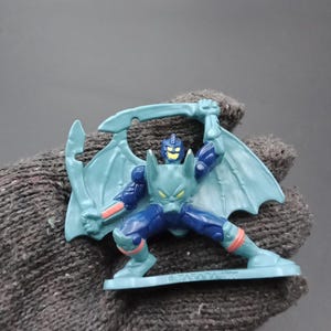 May include: A blue and navy action figure of a winged creature holding a sword. The figure has orange accents and is posed in a dynamic stance. The figure is on a small, rectangular base.