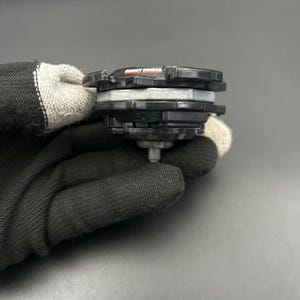 May include: A black and gray toy with the text "MIGHT DRIGER" in orange. The toy has multiple layers and a silver metal tip. The toy is held by a person wearing a black glove.