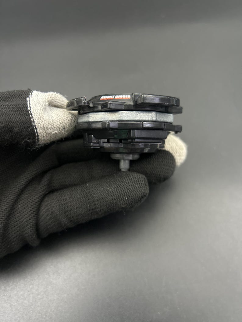 May include: A black and gray toy with the text "MIGHT DRIGER" in orange. The toy has multiple layers and a silver metal tip. The toy is held by a person wearing a black glove.