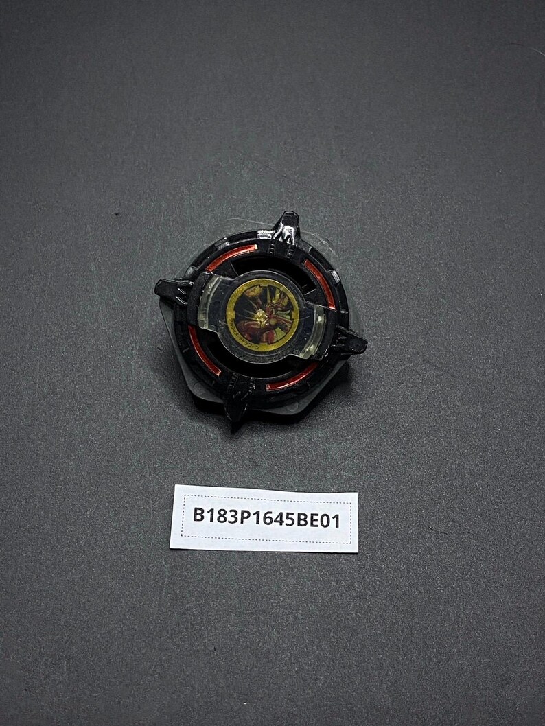 K&ouml;nnte beinhalten: Schwarzes und rotes Kreisel mit goldener Mitte und einem schwarz-wei&szlig;en Design. Der Kreisel hat einen Aufkleber mit dem Text "B183P1645BE01".