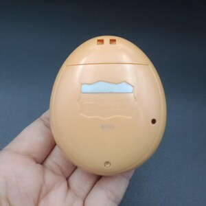 May include: A light orange, egg-shaped handheld electronic device. The device has a small rectangular screen and two small square buttons at the top. The back of the device has a small circular button and a small rectangular opening.