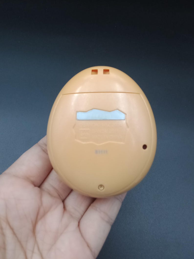 May include: A light orange, egg-shaped handheld electronic device. The device has a small rectangular screen and two small square buttons at the top. The back of the device has a small circular button and a small rectangular opening.