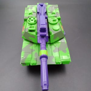 May include: A toy tank in shades of green and purple, featuring a camouflage pattern. The tank has a long purple barrel and a detailed design, suggesting a military theme. The toy is made of plastic.