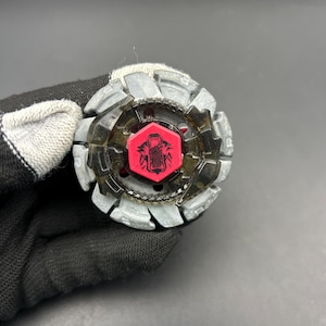 May include: A grey and black spinning top toy with a red centre. The top has a hexagonal red centre with a graphic design. The outer ring is grey with angular edges. The toy is designed for spinning and battling.