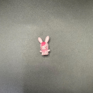 Puede incluir: Pequeña figura de conejo rosa con una raya rosa oscuro en la cabeza y un lazo rosa. El conejo tiene un diseño lindo y de dibujos animados y está de pie. El fondo es una superficie gris oscuro sólida.