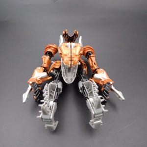 May include: A disassembled, metallic toy dinosaur with a detailed design. The toy features a combination of orange, silver, and black components. The head and upper body are orange, while the legs and lower body are silver and black.