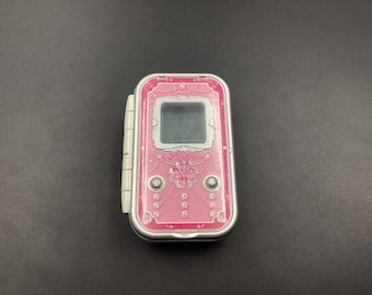 May include: A pink and silver handheld electronic game with a screen that displays "Pinky" and a flower design. The game has a silver border and a white button on the side.