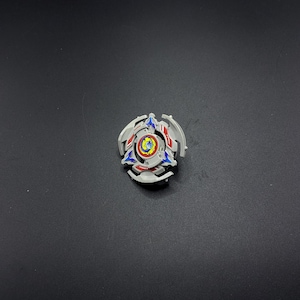 Könnte beinhalten: Ein weißer, blauer und roter Beyblade mit einem gelben und blauen Zentrum. Der Beyblade hat ein sternförmiges Design auf der Oberseite.