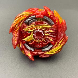 Könnte beinhalten: Ein kreisförmiges Kreiselspielzeug in leuchtendem Rot und Gold. Das Design umfasst komplexe, flammenartige Vorsprünge, die vom Zentrum ausgehen, sowie metallisch silberne Akzente und ein kleines grünes Detail auf der zentralen Vorderseite.