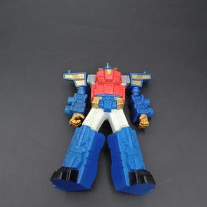 May include: A colorful toy robot with a red chest, blue arms and legs, and gold accents. The toy has a white midsection and black feet. The robot is standing upright against a dark gray background.