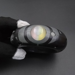 May include: Black and grey oval device with a central, glowing, multi-coloured light. The device has a series of grey curved lines and a small button on the side. The object is held by a person wearing a black glove.