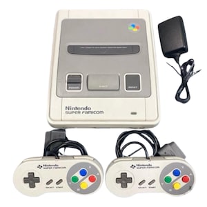 Könnte beinhalten: Eine hellgraue Nintendo Super Famicom Spielkonsole mit dunkelgrauem Kassettenschacht sowie Power-, Eject- und Reset-Tasten. Enthält zwei hellgraue Controller mit Steuerkreuz, Select-, Start-Tasten und vier farbigen Aktionstasten sowie ein schwarzes Netzteil.