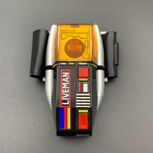 May include: A black, silver, and orange plastic toy device featuring the word LIVEMAN printed vertically in white letters. The device includes a translucent orange top section, a central red button, and vertical blue and yellow stripes on the side.