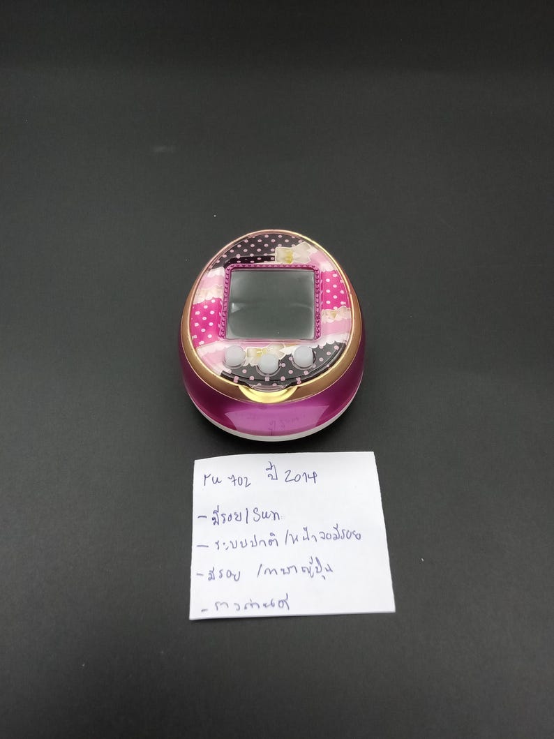 Puede incluir: Un juego electr&oacute;nico port&aacute;til rosa y dorado con pantalla. La secci&oacute;n superior presenta un patr&oacute;n floral y de lunares. El dispositivo tiene tres botones debajo de la pantalla. Hay una nota manuscrita al lado del dispositivo.