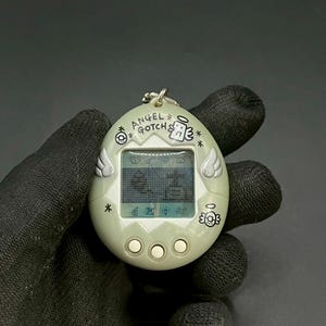 Könnte beinhalten: Ein hellgrünes Angel Gotch Virtual Pet Gerät. Das eiförmige Gerät hat einen kleinen Bildschirm, der pixelige Grafiken anzeigt. Das Gerät hat drei Knöpfe unter dem Bildschirm und Engelsflügel-Designs an den Seiten. Der Text "ANGEL GOTCH" ist oben aufgedruckt.