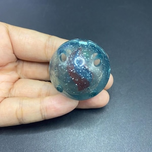 Puede incluir: Un objeto esférico transparente con un tono azul-verdoso y detalles internos rojos y blancos. La esfera tiene dos pequeños agujeros y un aspecto moteado y brillante. El objeto se sostiene en una mano sobre un fondo oscuro.