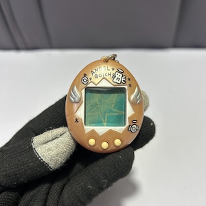 May include: A vintage, tan-colored Angel Gotcha electronic pet game. The handheld device features a small screen displaying a star graphic, with three buttons below. The casing has angel wing and skull details, with the text "ANGEL GOTCHA" visible.
