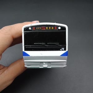 Puede incluir: Un modelo de tren en miniatura con una combinación de colores blanco, plateado y azul. La parte frontal del tren presenta una ventana negra y una pantalla digital con texto japonés. El logotipo de JR es visible en el lateral.