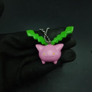 May include: A pink, cartoon-like keychain featuring a character with green leaf-like appendages. The character has small yellow eyes and a smiling mouth. The keychain is attached to a silver chain.