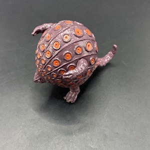May include: A purple, spherical figurine with orange circular details. The figurine has multiple tentacle-like appendages and a small, footed base. The overall design suggests a creature or monster-like aesthetic.