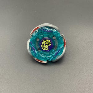 Pode incluir: Um brinquedo de pião circular com um anel exterior de metal prateado com detalhes em vermelho, uma secção central de plástico azul-petróleo translúcido com detalhes roxos e um parafuso hexagonal roxo com um ícone gráfico de dragão amarelo.