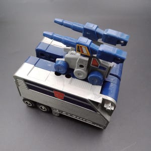 May include: A vintage toy vehicle in shades of blue, silver, and gray. The toy features a trailer-like base with wheels and a top section with two blue cannons. A red emblem is visible on the side.